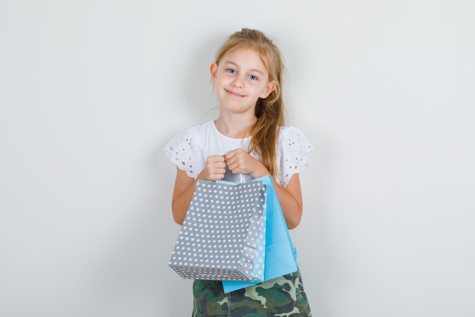 little girl holding paper bags in white t shirt, skirt and looking happy. front view.